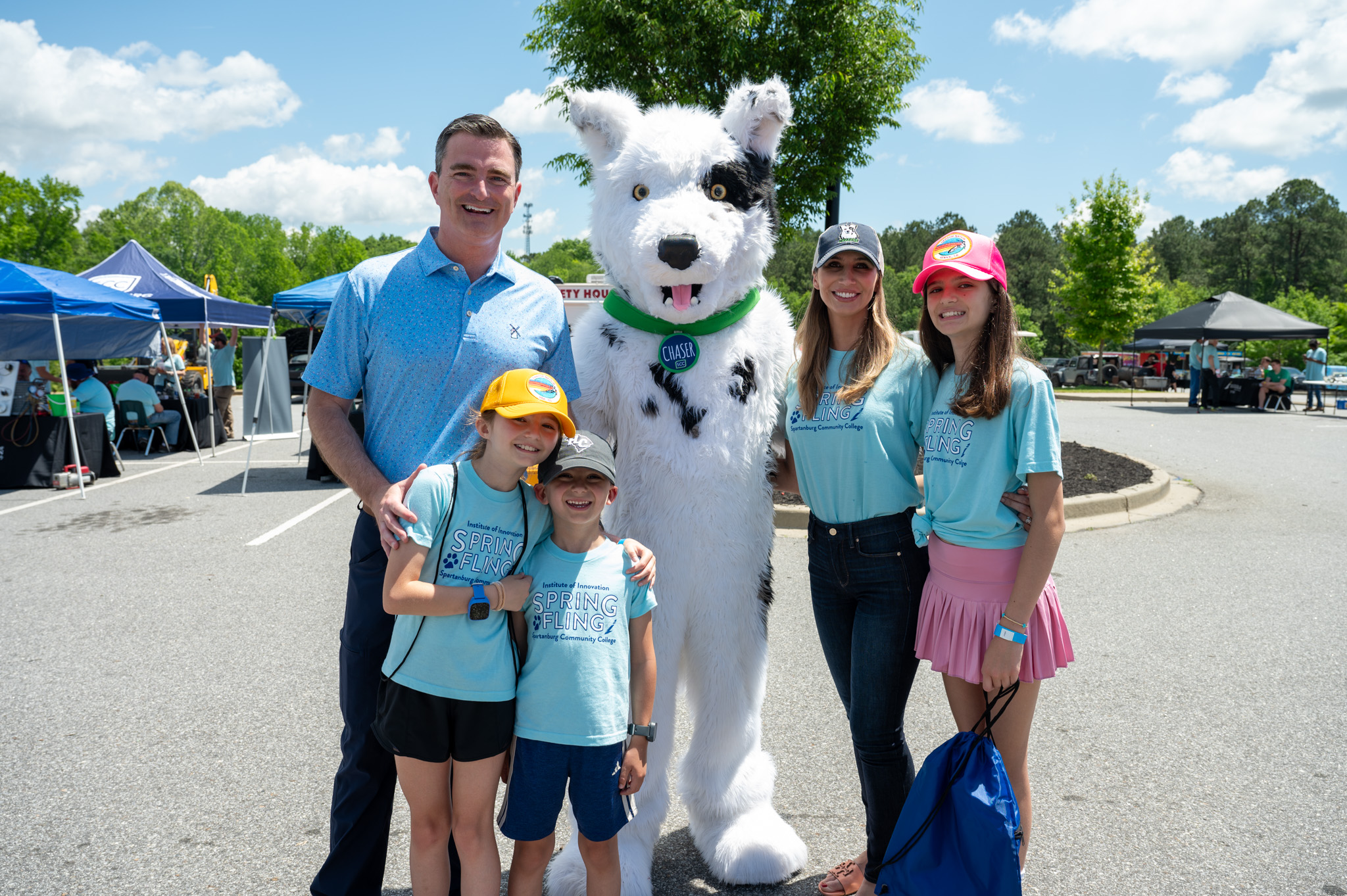 A group of five people and a large white dog mascot pose together outdoors at a sunny community event, with tents and booths in the background. Two children in matching blue “Spring Fling” shirts stand in front, while three adults and the mascot stand behind them, all smiling for the photo.