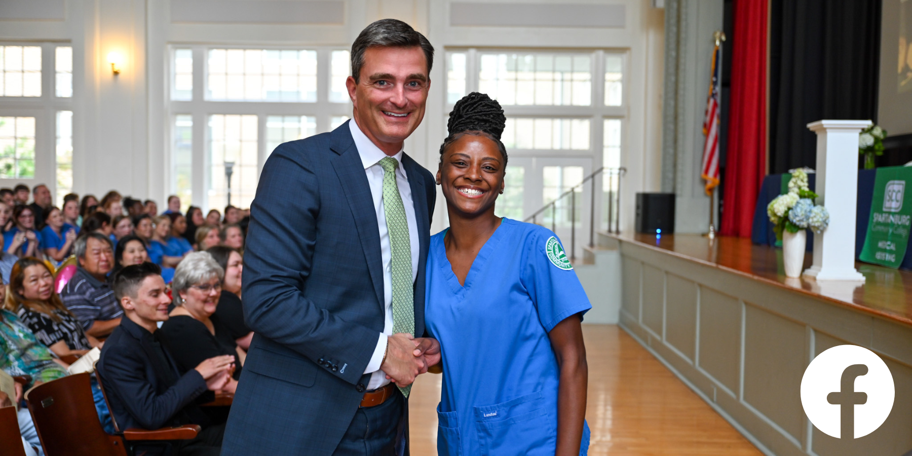 Individual in business attire shakes hands with an individual wearing blue medical scrubs at the front of an indoor auditorium, with an audience seated behind them and a stage featuring an American flag and SCC signage, capturing a formal recognition or award moment in a campus setting.