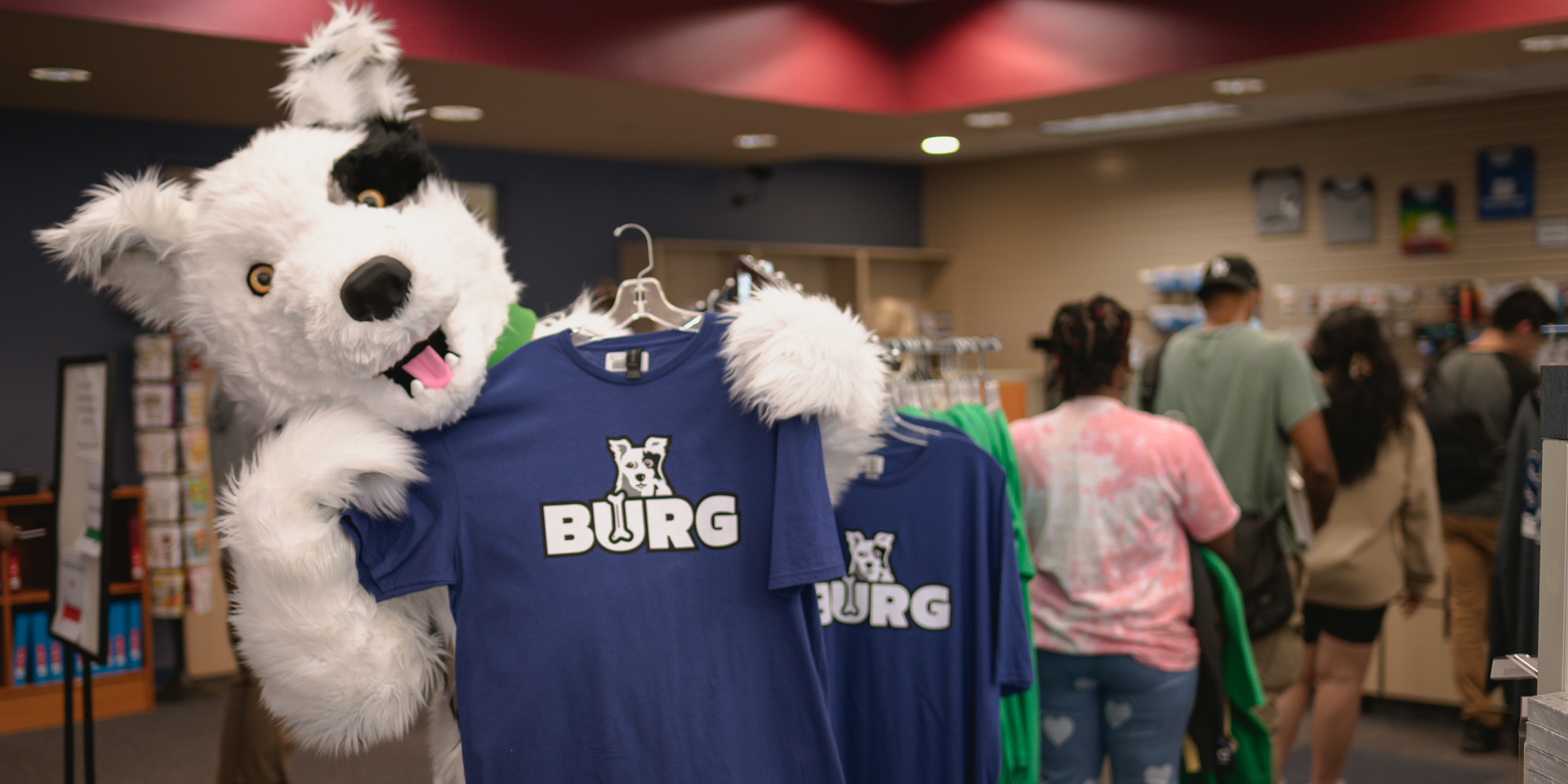 Chaser mascot from Spartanburg Community College stands inside a campus bookstore holding up a blue “BURG” t-shirt on a hanger, with additional shirts displayed on a rack. In the background, several people browse merchandise along shelves and clothing racks in a retail-style campus environment with warm lighting.