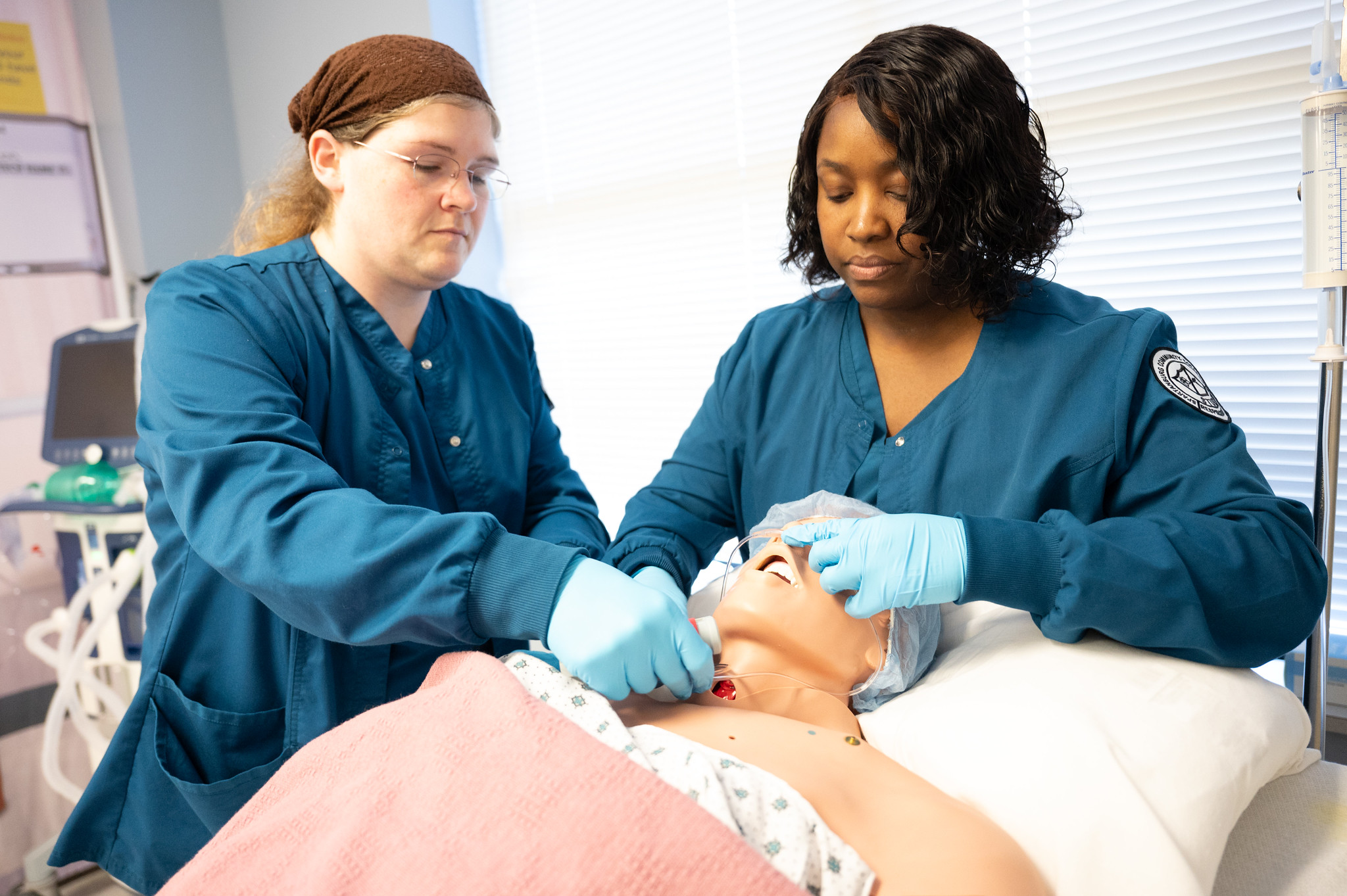 Two nursing students in teal scrubs practice medical procedures on a patient simulation mannequin in a training lab setting.
