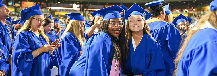 A wide, candid shot of two smiling female graduates in bright blue caps and gowns looking at the camera, surrounded by other students in graduation attire at an outdoor stadium ceremony.