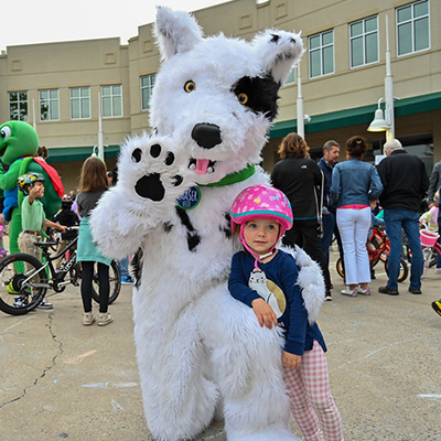 A large white mascot with a black eye patch and green collar poses and waves beside a young child wearing a pink helmet and patterned pants, standing together outdoors at a lively community event with people, bicycles, and a building in the background.