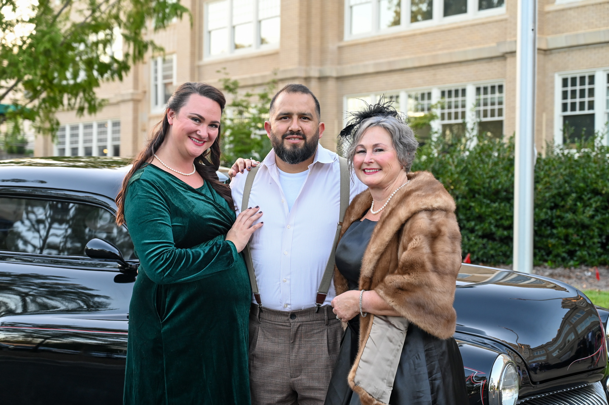 A group of three elegantly dressed guests pose in front of a vintage black car outside a historic building during a themed event. The women wear classic evening attire with pearls and fur, while the man sports suspenders and a white shirt, evoking a timeless, 1940s-inspired atmosphere.