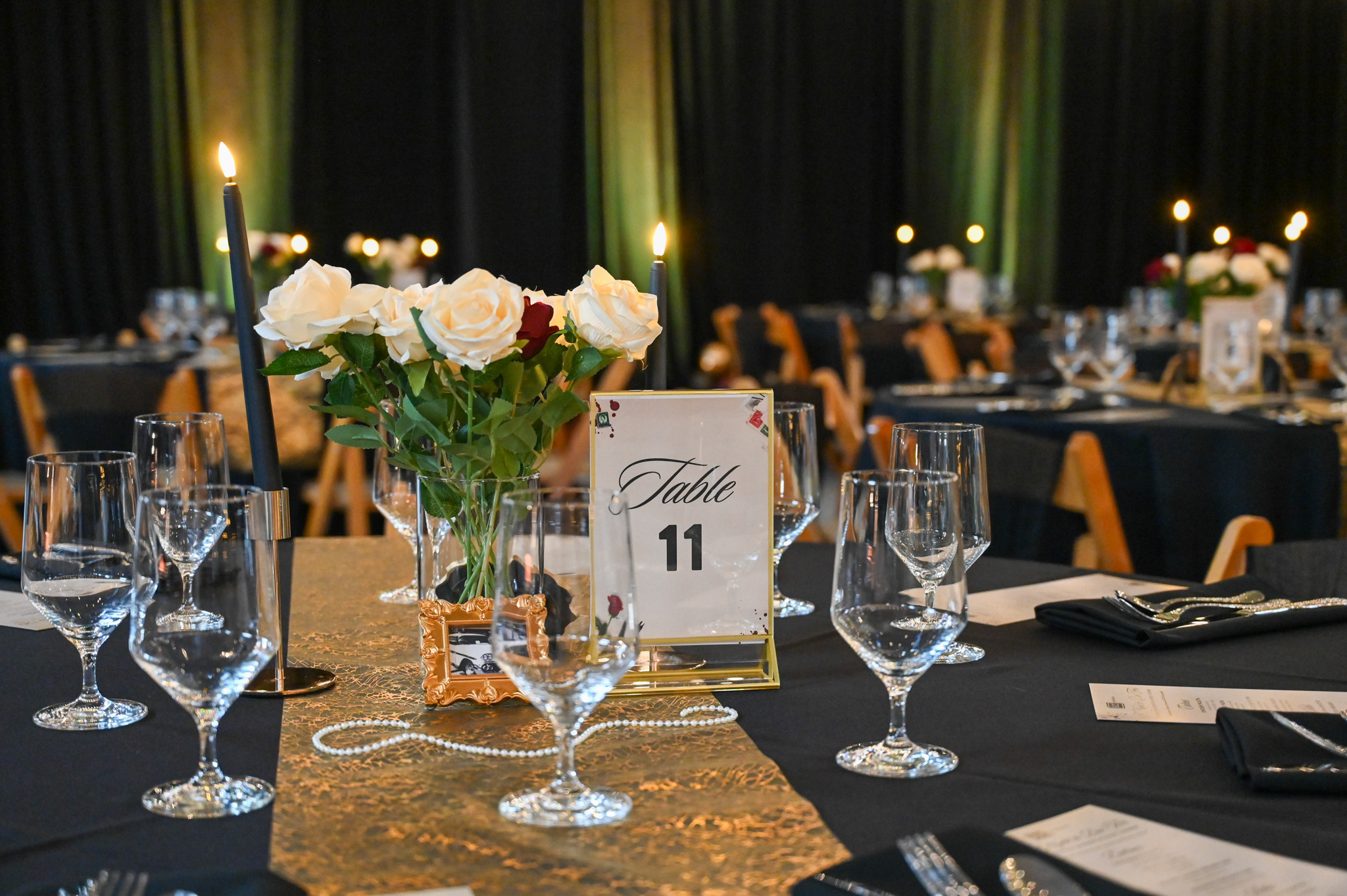 An elegant table featuring wine glasses, flowers, a table number sign with eleven on it, and some fake pearls all on top of a black and gold table clothe