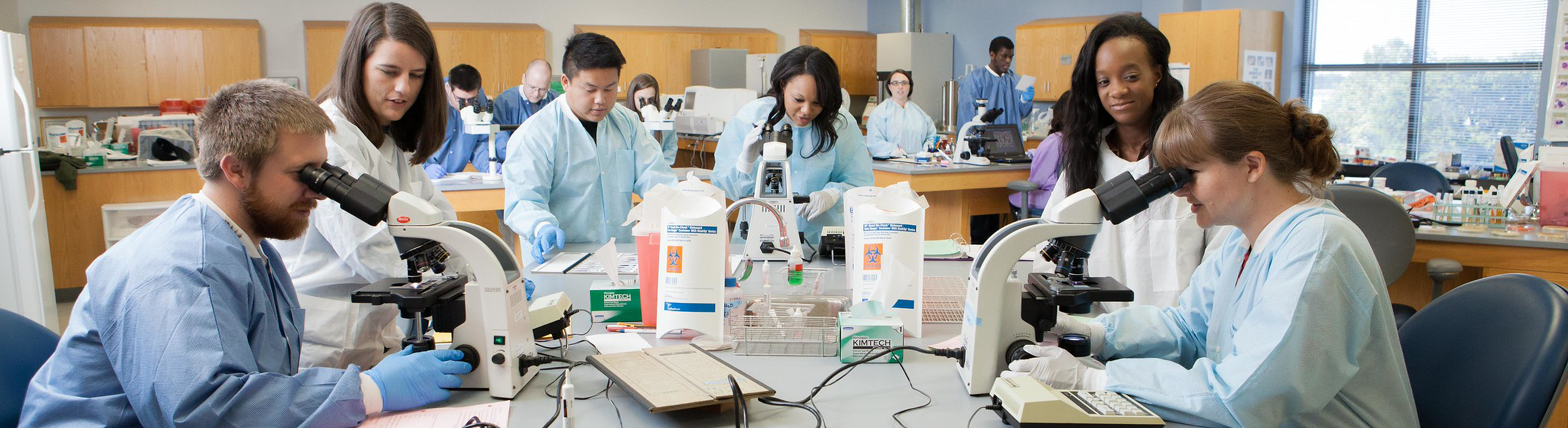 A group of students in a science laboratory wearing protective gowns and gloves work together at lab benches, using microscopes and handling samples while an instructor supervises in the background.