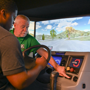 Two individuals seated inside a commercial truck driving simulator, with one in the driver’s seat holding the steering wheel while the other points to controls on the dashboard. A large screen displays a simulated roadway with trees, hills, and lanes ahead, indicating a hands-on CDL or driver training environment.