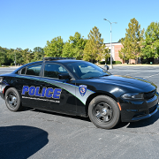 Side view of a black Spartanburg Community College police vehicle parked on a paved campus roadway, featuring large blue “POLICE” lettering and the SCC logo on the door. Trees, a campus building, and a clear blue sky are visible in the background.