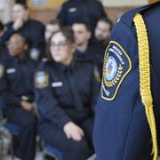 Close-up view of a public safety uniform displaying a shoulder patch and ceremonial cord in the foreground, with multiple uniformed students seated in rows in the background. The indoor setting appears to be a law enforcement or public safety training ceremony, with participants dressed in matching academy-style uniforms.