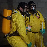 Two individuals wearing yellow hazmat suits, protective gloves, and full-face respirators stand indoors handling equipment in a controlled environment. One holds a device while the other stands nearby with a large tank secured on the back, indicating hazardous materials or emergency response training.