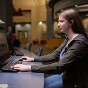 Individual seated at a desktop computer workstation inside a campus lab types on a keyboard while focusing on a monitor. The setting includes multiple computers arranged along a counter, with chairs, a softly lit interior, and additional workstations visible in the background, indicating a student technology or study environment.