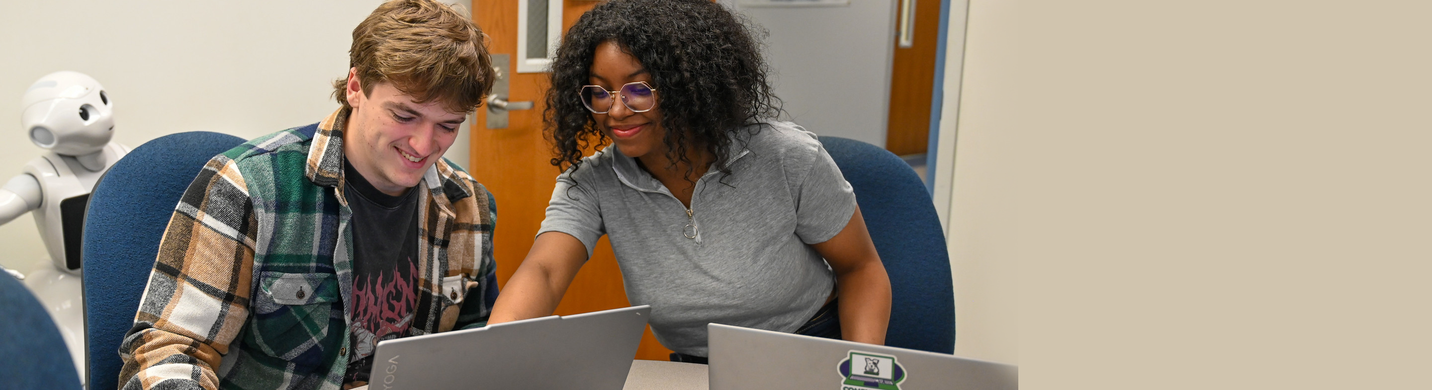 Two students collaborate on laptops at a table, smiling as they review work together, with a small robot visible in the background.
