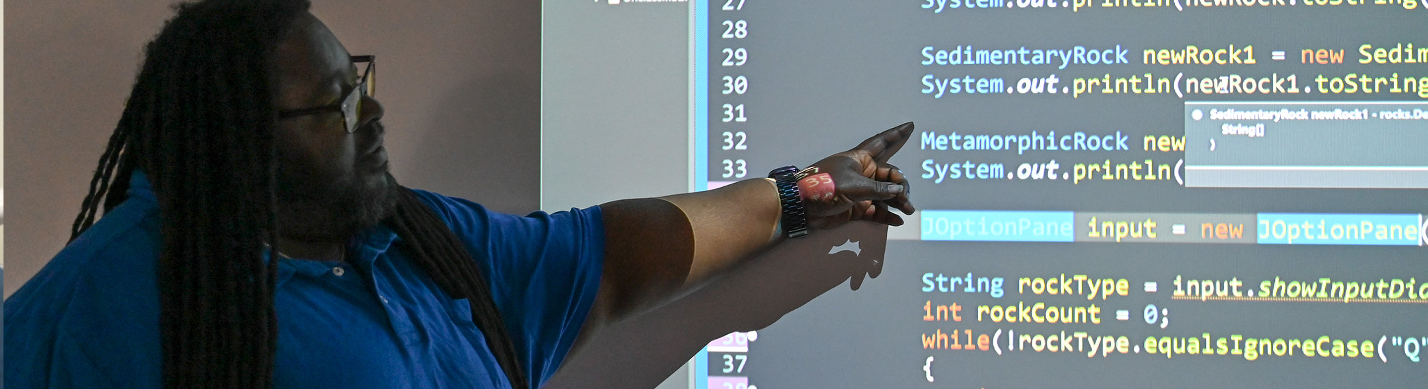 A man with long dark hair and a blue shirt points at a large projector screen displaying lines of Java programming code. The code mentions rock types like SedimentaryRock and MetamorphicRock, as well as a while loop for user input.