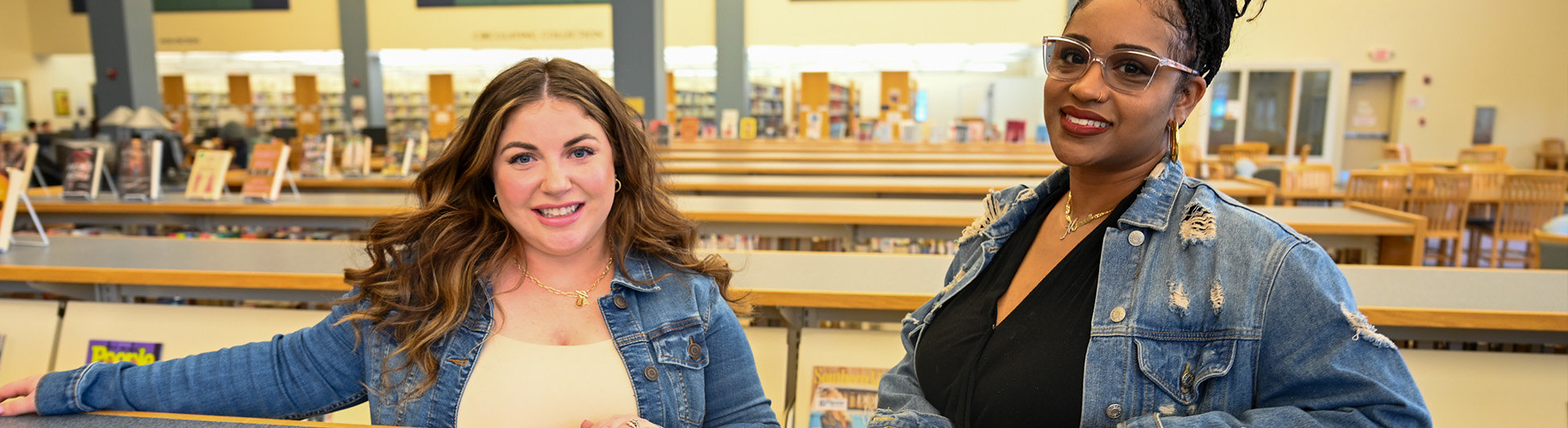Two women standing and smiling at a counter inside a library, both wearing denim jackets, with bookshelves and reading tables visible in the background.