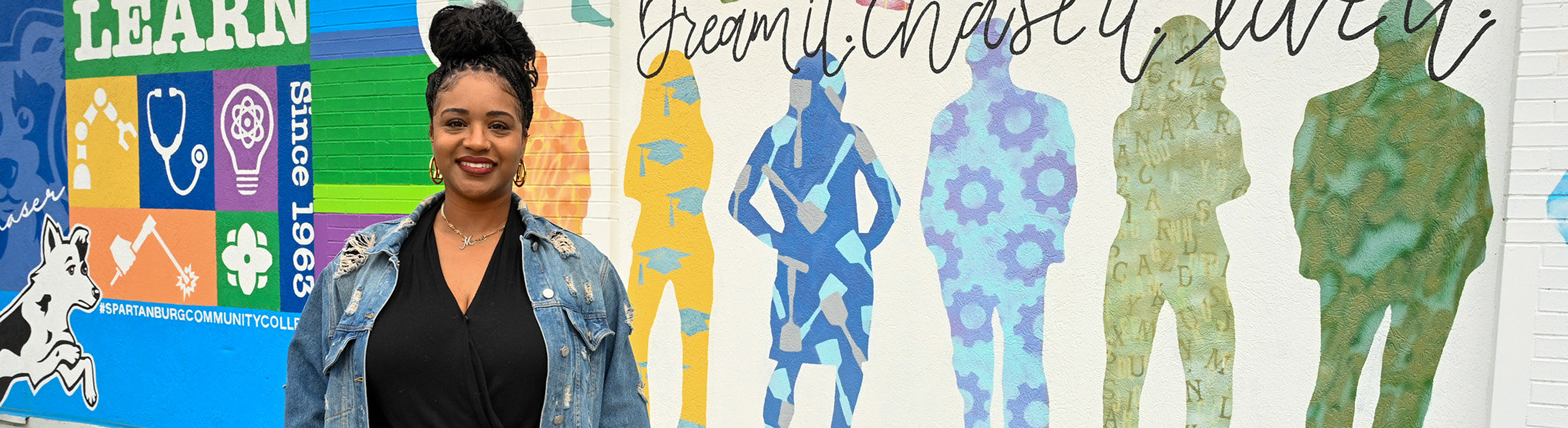 Smiling person outdoors wearing jean jacket, a black shirt, standing on a campus walkway with greenery and buildings in the background.