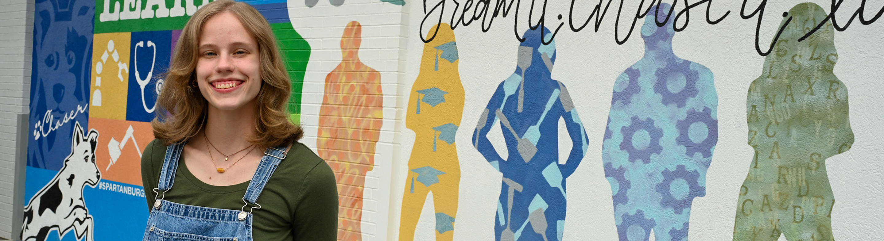 Smiling person outdoors, wearing jean overalls, a green shirt, and a gold necklace, standing on a campus walkway with a mural in the background.