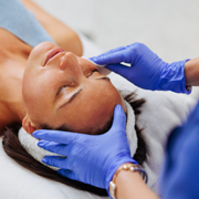 Close-up view of an esthetics procedure where a practitioner wearing blue gloves applies a treatment to a client’s eyebrow area using a small brush. The client lies on a treatment bed with eyes closed while the practitioner steadies the forehead, indicating a facial or eyebrow care service in a clinical or spa setting.