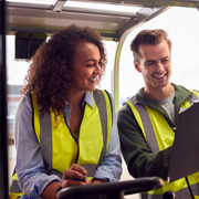 Two individuals wearing high-visibility safety vests stand inside a warehouse while looking at and interacting with a laptop placed on industrial equipment. The setting includes metal framework, storage areas, and industrial surroundings, indicating a logistics, manufacturing, or warehouse operations environment.