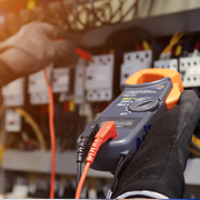 Close-up view of a technician wearing protective gloves using a digital multimeter to test connections inside an electrical control panel. The device is held in the foreground with red and black probes attached, while a panel filled with switches, relays, and wiring appears in the background, indicating electrical testing or maintenance work.