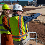 Two individuals wearing hard hats and high-visibility safety vests stand at a construction site, with one pointing toward an area under development while the other observes. The setting includes graded soil, construction zones, and equipment, indicating supervision or project coordination activity.