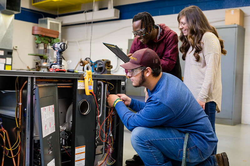 A diverse group of five students is gathered in a brightly lit technical workshop, focused on a hands-on learning project involving an open HVAC unit. A student in the center, wearing a maroon and black windbreaker and clear safety glasses, smiles while carefully working with wires inside the machinery. To his left, another student in a brown cap and safety goggles holds a yellow multimeter