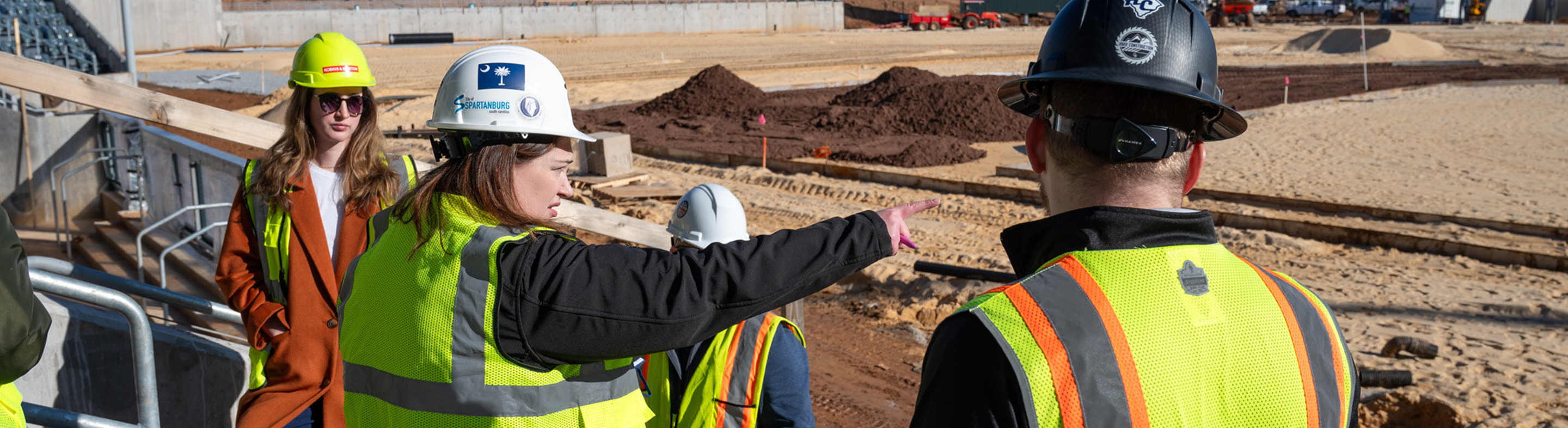 Four individuals wearing hard hats and high-visibility safety vests stand together at a large construction site, with one pointing toward an area under development. The scene includes graded dirt, construction materials, equipment, and marked zones, indicating a collaborative construction supervision or project planning environment.