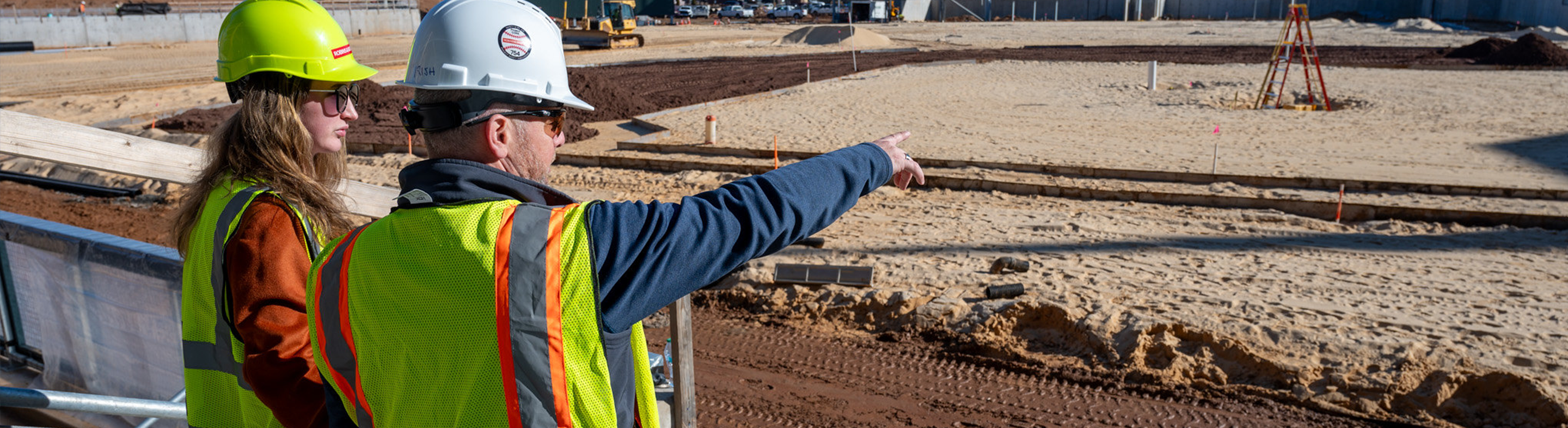 wo individuals wearing construction hard hats and high-visibility safety vests stand at an active job site, with one pointing toward an area under development while the other looks on. The scene includes dirt grading, heavy machinery, measuring equipment, and marked construction zones, indicating a construction supervision or project coordination environment.