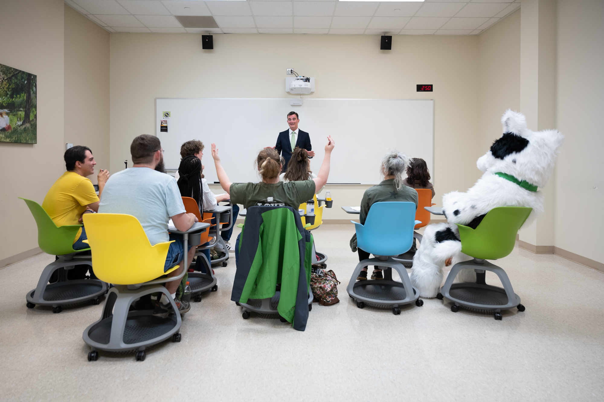 Instructor stands at the front of a classroom speaking while students sit in colorful rolling chairs facing forward, with several raising their hands to participate. A person in a large dog mascot costume sits among the students, and the room includes a whiteboard, wall clock displaying 2:50, speakers, and classroom walls with a framed picture.