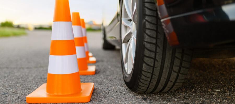 Close-up view of a car’s tire positioned next to a row of orange traffic cones on a paved roadway. The cones are evenly spaced, and the scene focuses on the tire and cones alignment, indicating a controlled driving scenario such as a defensive driving course or vehicle training exercise.