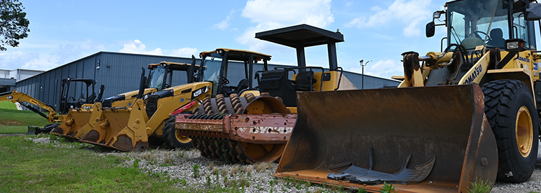Row of heavy construction equipment parked outdoors on a gravel and grass surface, including bulldozers, compactors, and loaders with large metal blades and rollers in the foreground. A warehouse-style building and blue sky with scattered clouds are visible in the background, indicating a heavy equipment training or industrial setting.
