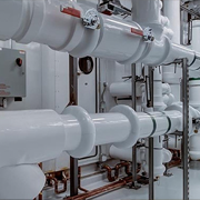A mechanical room filled with complex industrial HVAC piping systems. Large, insulated white pipes run horizontally and vertically across the room. They are supported by metal struts and brackets. An electrical control box sits on the wall behind the pipes. The entire room has a sterile white floor and ceiling.