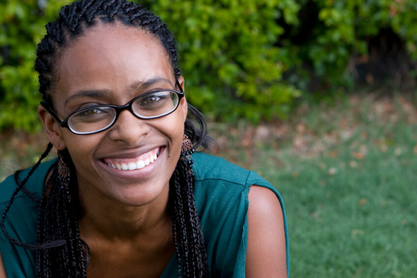 Close-up outdoor portrait of an individual wearing glasses and a green sleeveless top, positioned in front of a softly blurred background of grass and leafy greenery, with natural lighting highlighting facial detail and creating a clean, approachable campus-style headshot image.