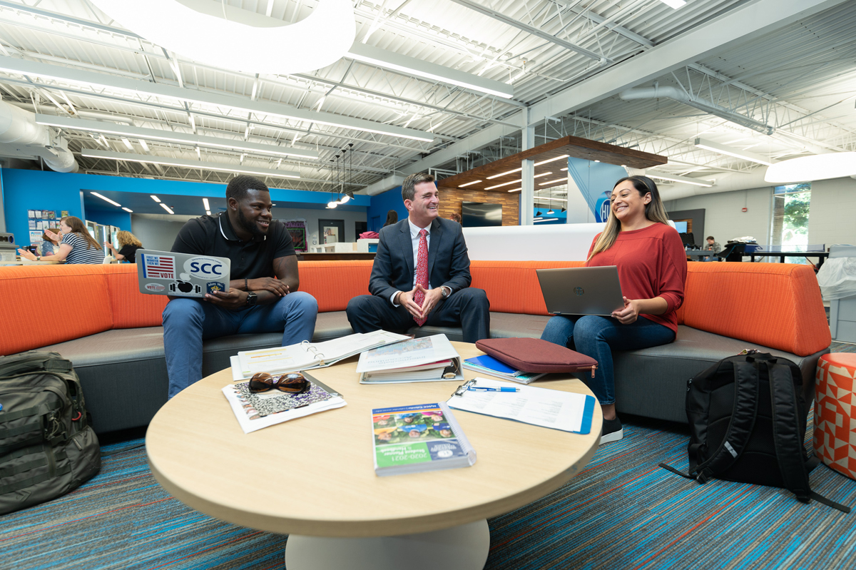 Modern campus lounge at Spartanburg Community College featuring three individuals seated on an orange curved bench around a round table with notebooks, folders, and books, using laptops and discussing materials, with a bright open interior showing exposed ceiling beams, large windows, and collaborative study space design.