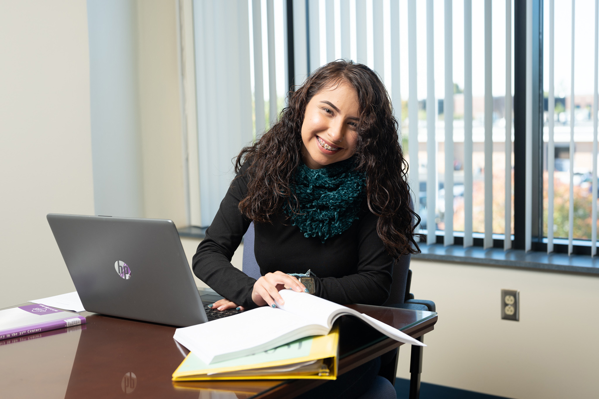 Student seated at a desk inside a bright classroom, working with an open book and laptop while other textbooks and folders are stacked nearby, with large windows and vertical blinds in the background allowing natural light into a clean academic study environment.