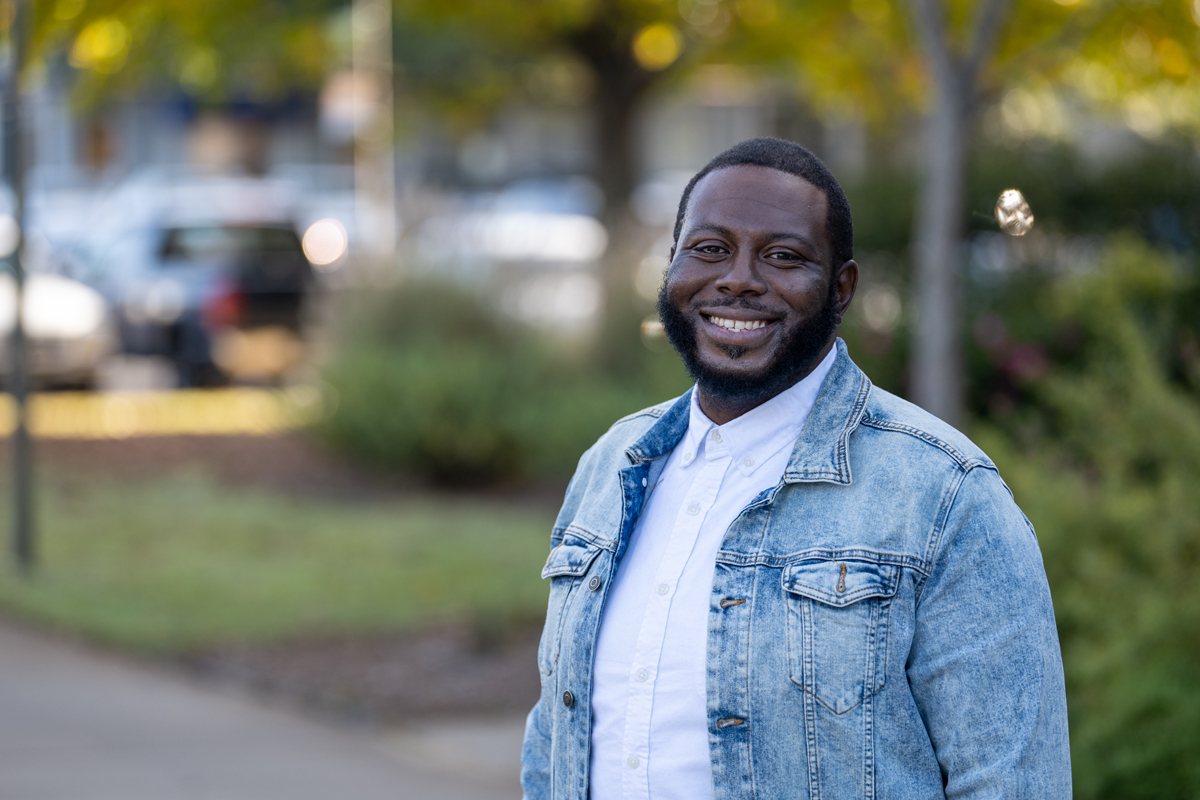 Outdoor portrait of an individual standing along a tree-lined campus walkway, wearing a light denim jacket over a white button-up shirt, with a softly blurred background of greenery, parked cars, and sunlight, creating a clean and professional college lifestyle image.