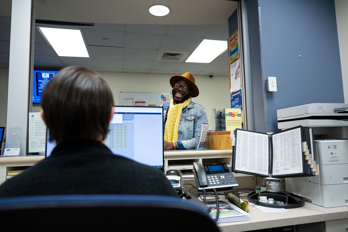 Interior student services office at Spartanburg Community College showing a front desk workstation with a computer, phone, and documents in the foreground, while an individual stands at the counter speaking with staff, with posters, signage, and office equipment visible in a professional campus service environment.