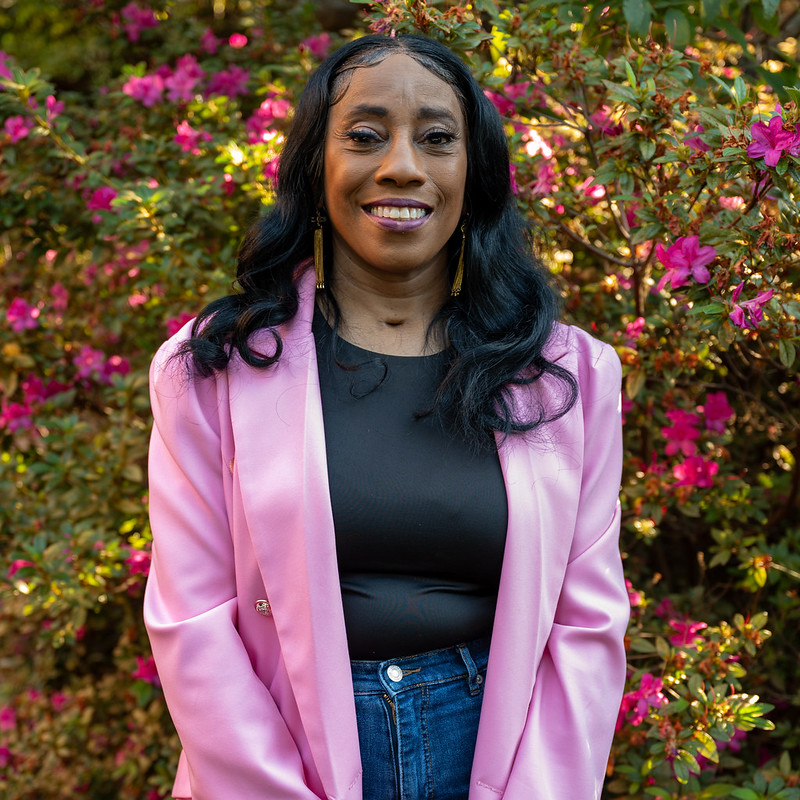 Outdoor portrait of an individual wearing a pink blazer over a dark top and jeans, standing in front of flowering shrubs with bright pink blossoms and dense greenery, with natural sunlight creating a softly blurred garden background for a clean professional campus-style headshot.
