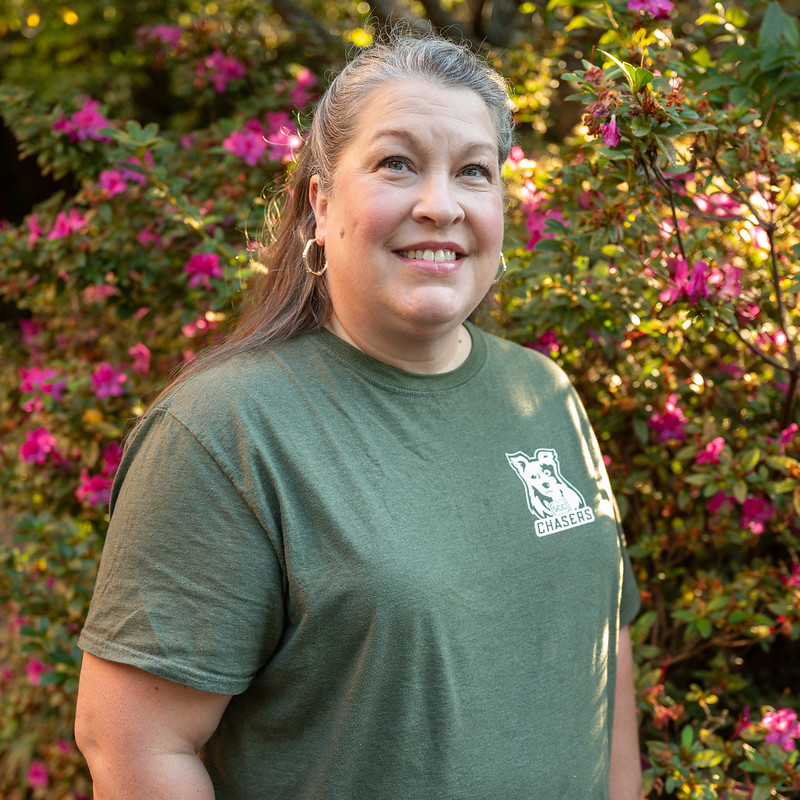 Outdoor portrait of an individual wearing a green Spartanburg Community College “Chasers” t-shirt, standing in front of flowering shrubs with pink blossoms and dense greenery, with natural sunlight creating warm highlights and a softly blurred garden background in a clean campus-style headshot.