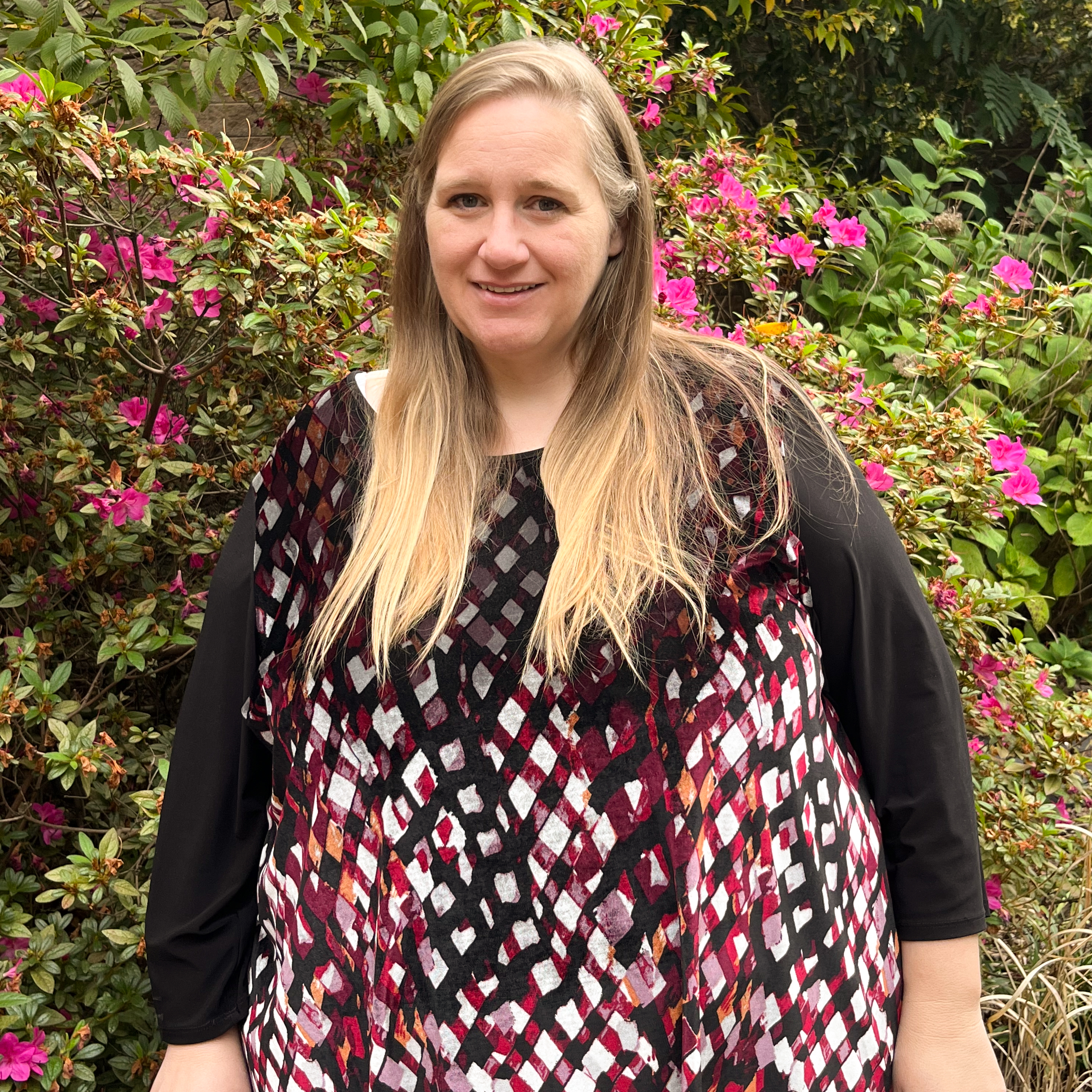 Outdoor portrait of an individual wearing a patterned top with red, black, and white geometric design and long sleeves, standing in front of flowering shrubs with pink blossoms and green foliage, with natural daylight and a softly blurred garden background creating a clean, professional campus-style headshot image.