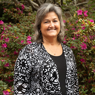 Outdoor portrait of an individual wearing a black patterned jacket over a dark top, standing in front of flowering shrubs with pink blossoms and dense green foliage, with soft natural lighting and a blurred garden background creating a clean, professional headshot-style image.