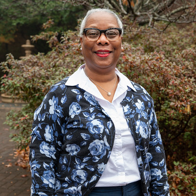 Outdoor portrait of an individual wearing glasses and a floral-patterned jacket over a white collared shirt, standing in front of dense shrubs and greenery with a garden feature partially visible in the background, captured in soft natural lighting with a clean, professional campus-style headshot composition.