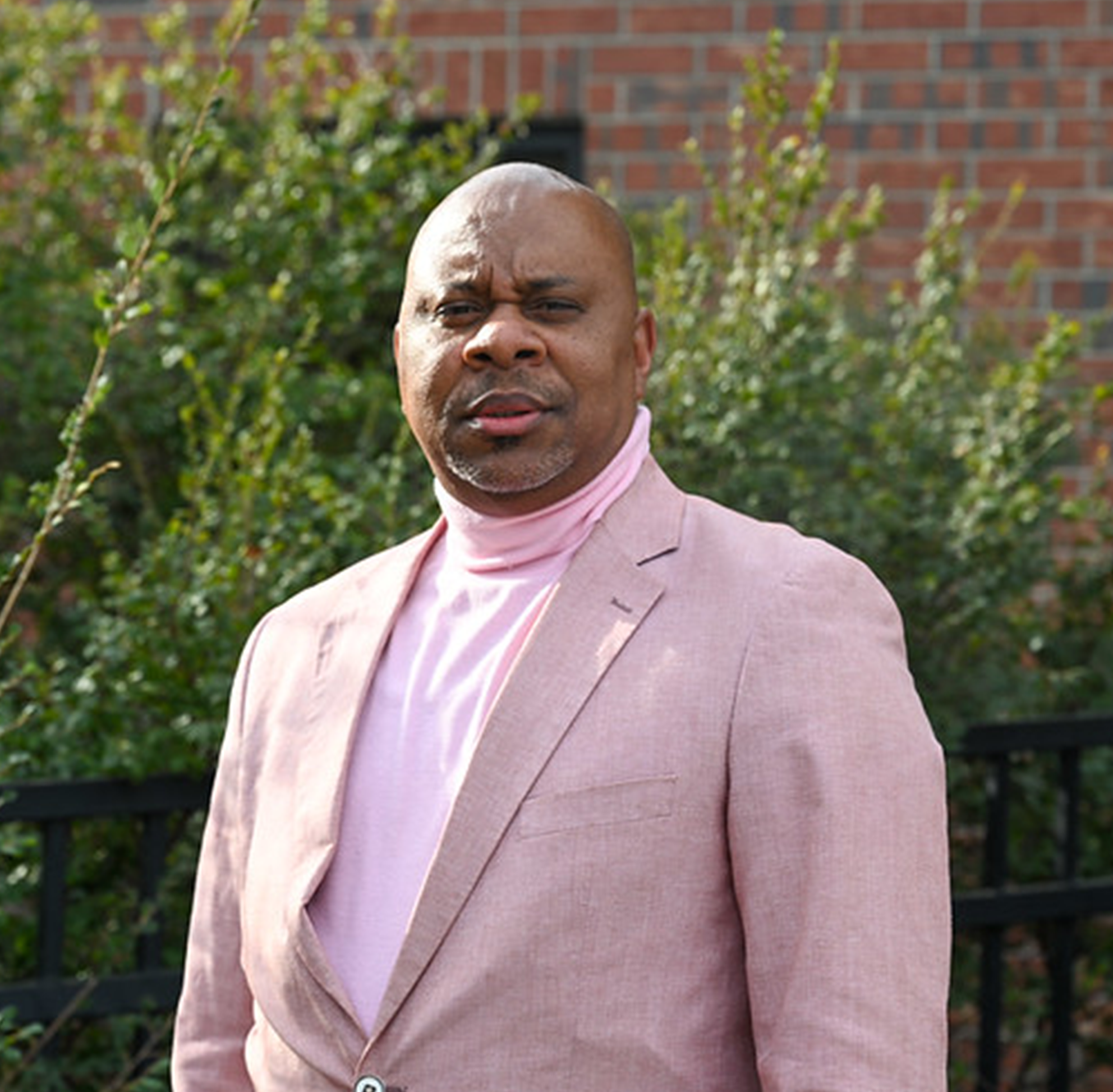 Outdoor portrait of an individual wearing a light pink blazer over a matching turtleneck, standing in front of green shrubs and a brick building, with natural sunlight creating a softly blurred background, resulting in a clean and polished professional headshot-style campus image.