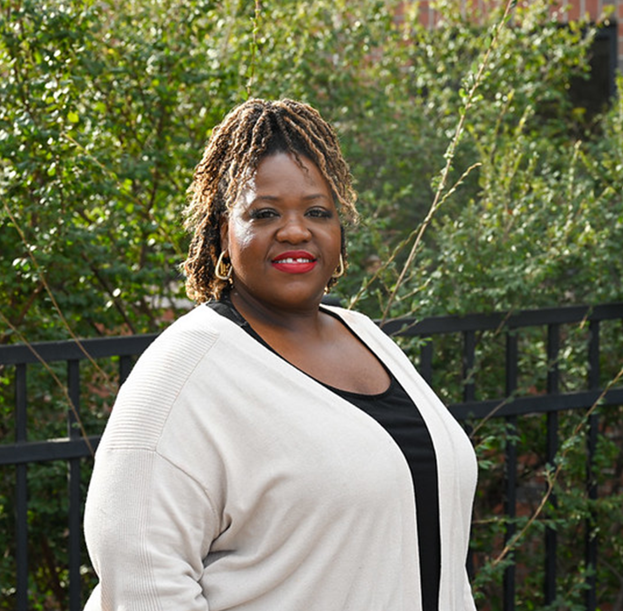 Outdoor portrait of an individual wearing a light cardigan over a dark top, standing in front of green shrubs and a brick wall, with textured hair styled upward and earrings visible, captured in bright natural daylight with a softly blurred background, creating a clean and professional headshot-style campus image.
