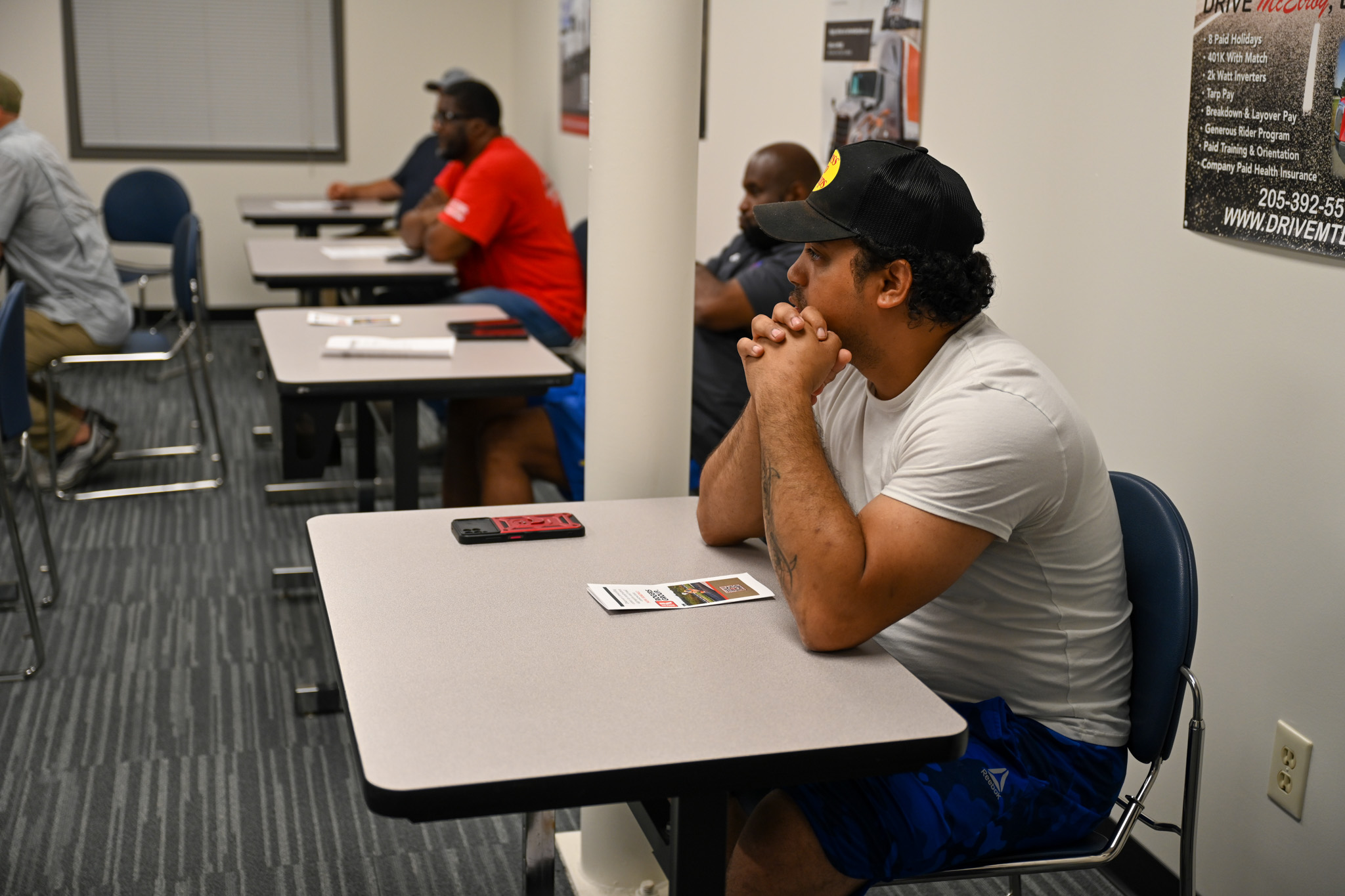 Adult learners seated at desks inside a classroom training environment, with one individual in the foreground resting hands together while facing forward, and several others seated behind at tables with notebooks and materials, surrounded by posters and signage on the walls, creating a focused workforce training or continuing education setting.