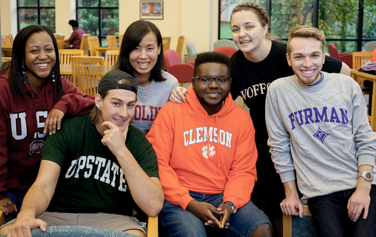 Group of six students seated and standing together in a classroom or study space with wooden tables and chairs, wearing college-branded apparel including USC, Clemson, Furman, Wofford, and Upstate, arranged closely for a group portrait with large windows and greenery visible in the background.