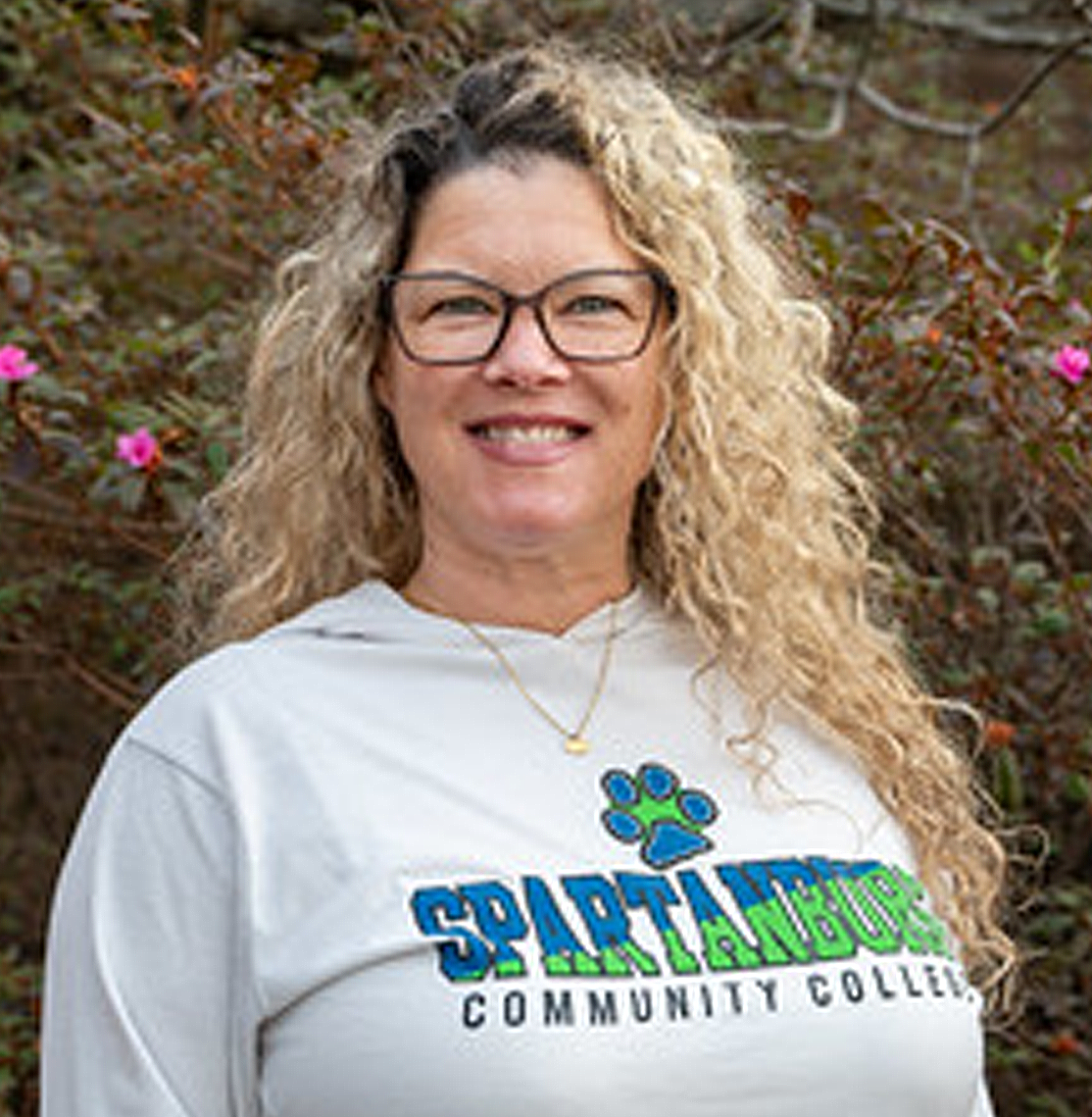 Outdoor portrait of an individual wearing glasses and a gray hoodie with “Spartanburg Community College” text and paw logo printed on the front, standing in front of leafy shrubs with small pink flowers, with natural light and a softly blurred background creating a clean, professional campus-style image.