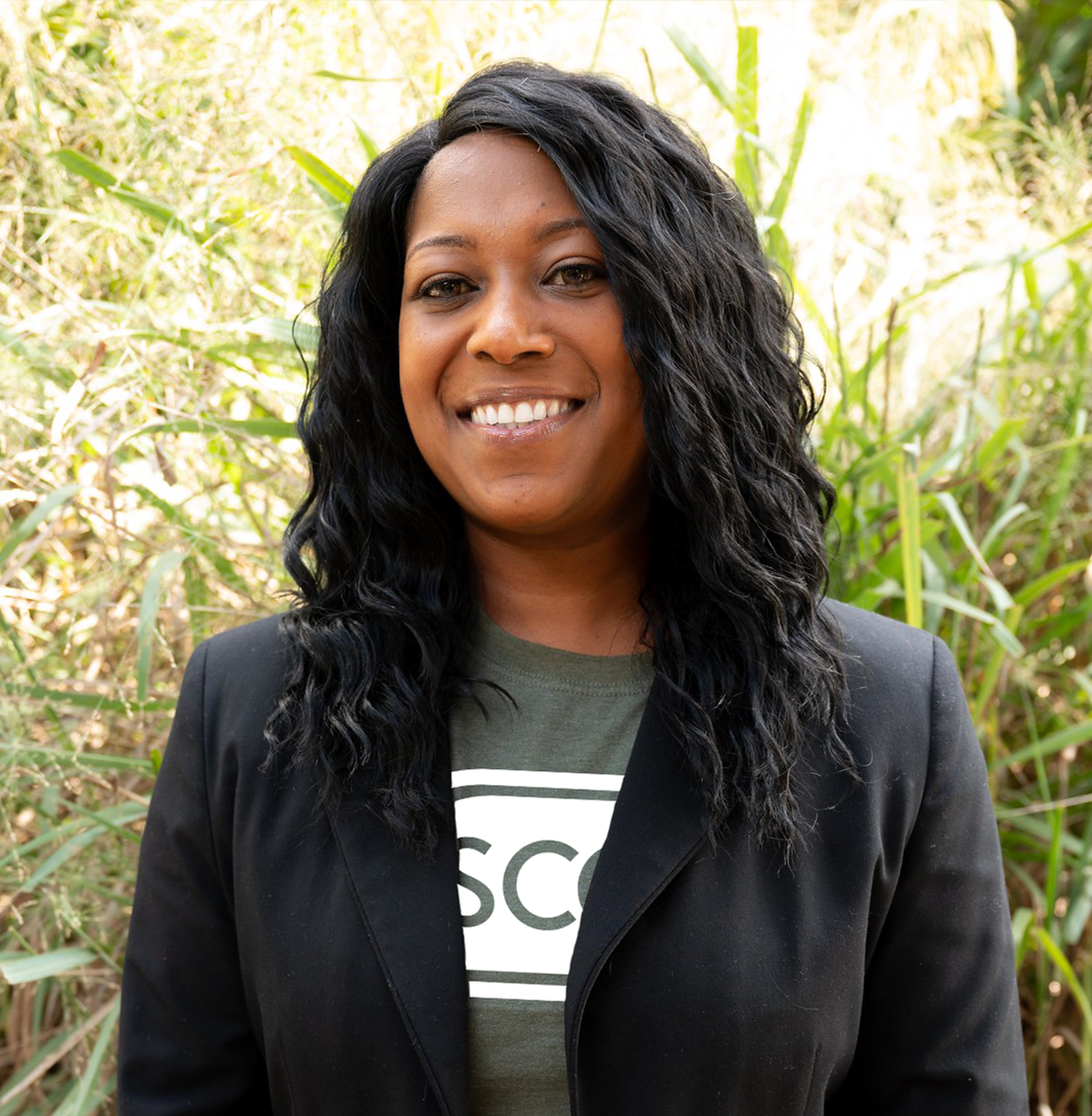 Outdoor portrait of an individual standing in front of tall green grasses and foliage, wearing a black blazer over a Spartanburg Community College shirt with visible “SCC” lettering, paired with dark jeans and a wristwatch, with natural daylight creating a softly blurred background and a clean, professional campus-style headshot composition.