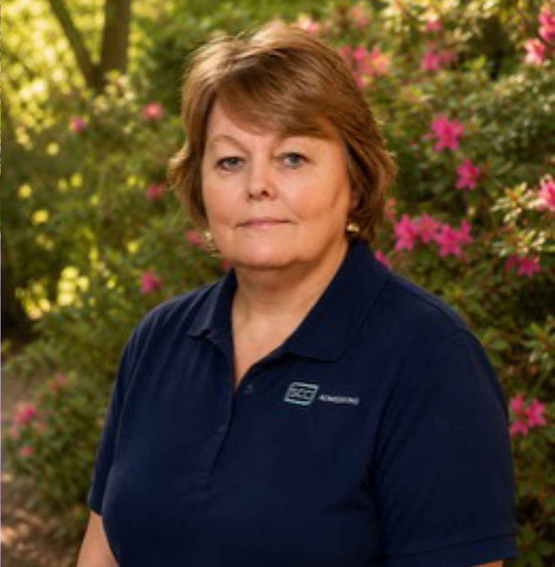 This image features a medium close-up portrait of a middle-aged woman with short, styled light brown hair and fair skin. She is looking directly forward with a neutral expression, wearing a navy blue polo shirt that features a small logo on the left chest containing the letters 