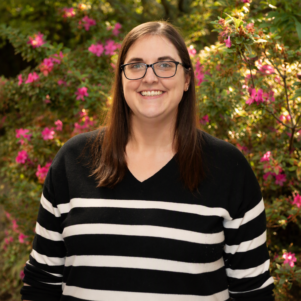 Professional outdoor headshot of a smiling woman with glasses and long brown hair, wearing a black-and-white striped sweater, standing in front of blooming pink flowers and lush greenery in natural sunlight.