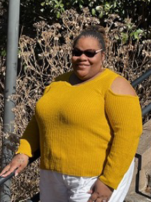 A person wearing a mustard yellow cold-shoulder sweater and white pants stands outdoors near a staircase railing, smiling while wearing sunglasses. Shrubs and greenery are visible in the background.