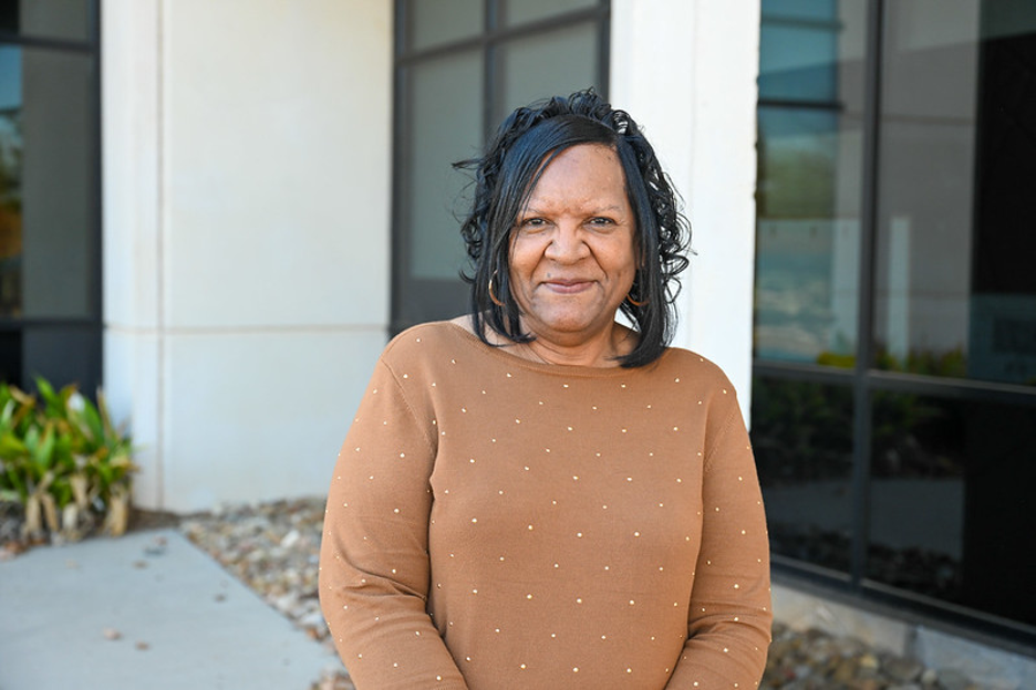 A woman stands outside a building, smiling at the camera. She is wearing a light brown long-sleeve top with small decorative dots. Behind her are large windows, a light-colored wall, and some landscaping with plants and stones.